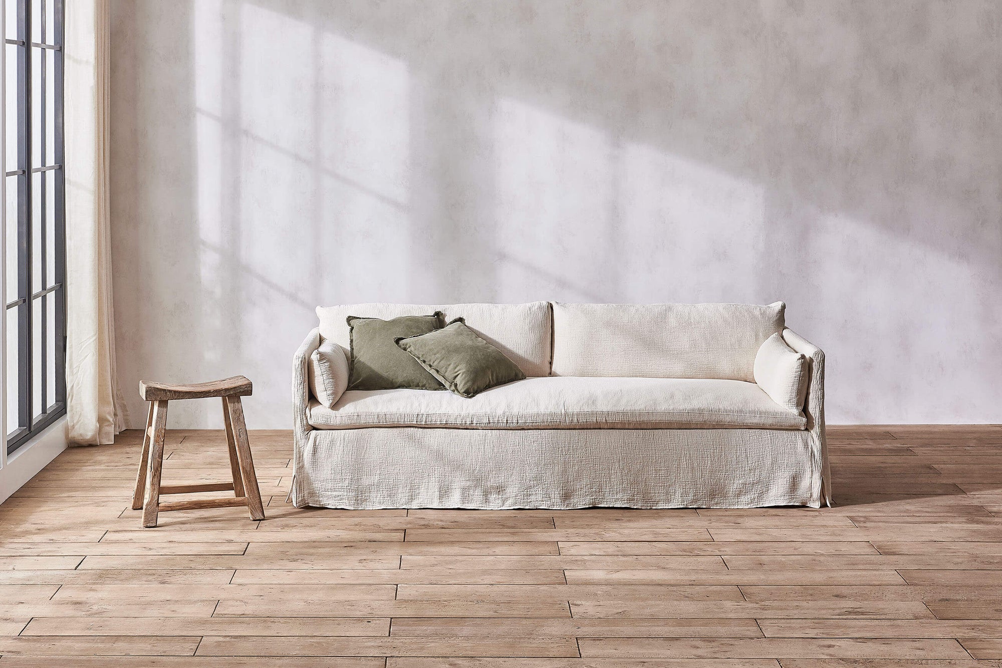 Gabriel 84" Sofa in Corn Silk, a light beige Washed Cotton Linen, with two sage throw pillows, next to a wooden stool