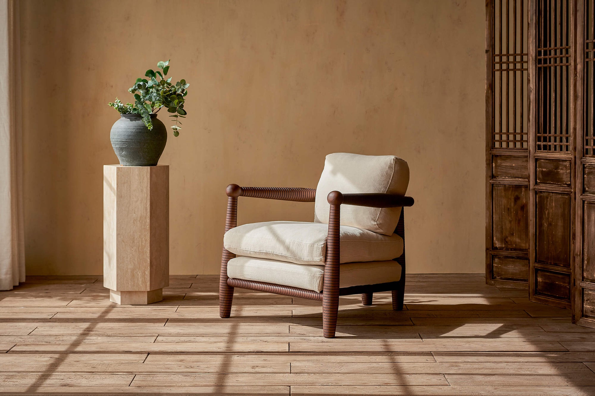 Gio Chair in a Dusky Ash wood frame with Washed Cotton Linen cushions in Corn Silk, placed in a sunlit room next to a potted plant on top of a decorative pillar.