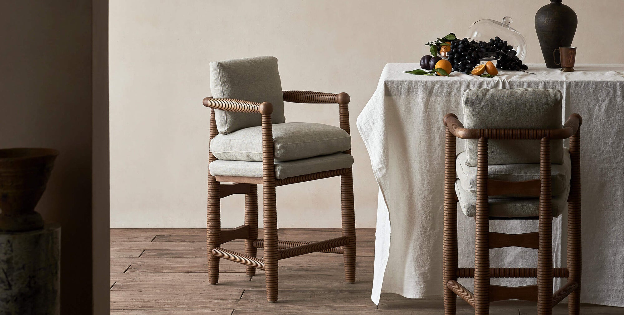 Two Gio Dining Stools in Heritage Ash frame and cushions in Jasmine Rice, a light warm greige Medium Weight Linen, sitting at a counter height table covered in a white linen tablecloth