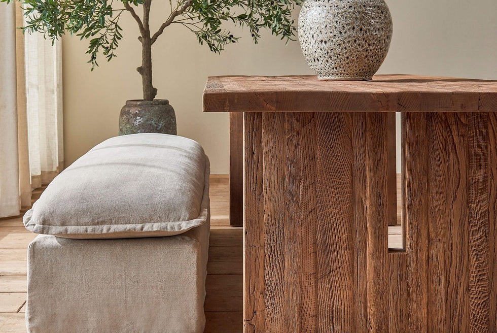 Close up of Kai Dining Table in 100% Reclaimed Austrian Heritage Oak, a medium, bosc pear shade with earthy undertones, placed in a sunlit room with a Neva Dining Bench and a potted tree