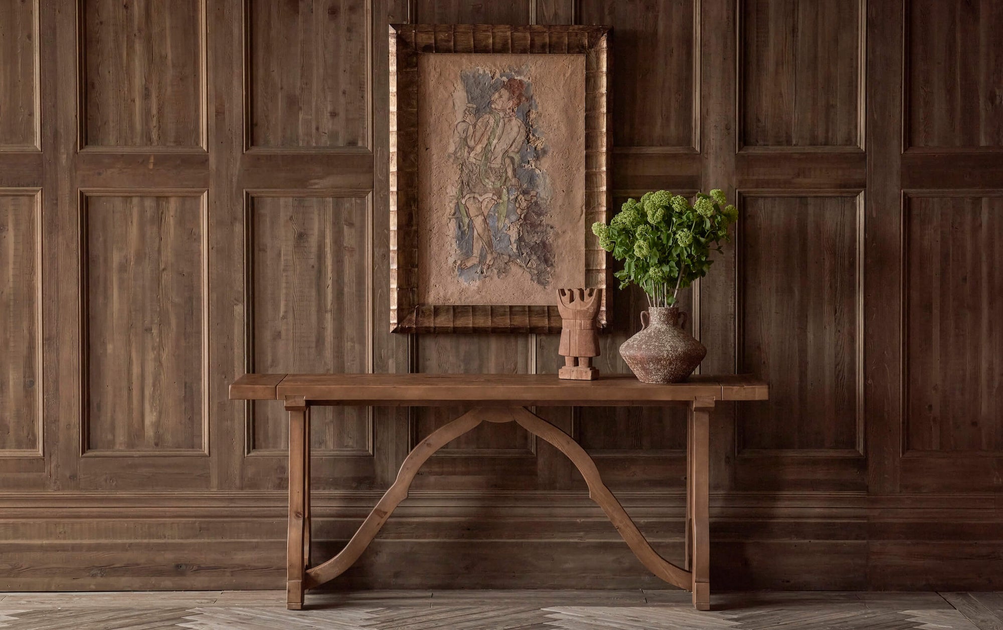 Leona Console Table in 100% Reclaimed Chinese Heritage Pine, a medium, bosc pear shade with earthy undertones, placed against a wood paneled wall, under a framed painting, and decorated with a wooden sculpture and a vase of flowers.