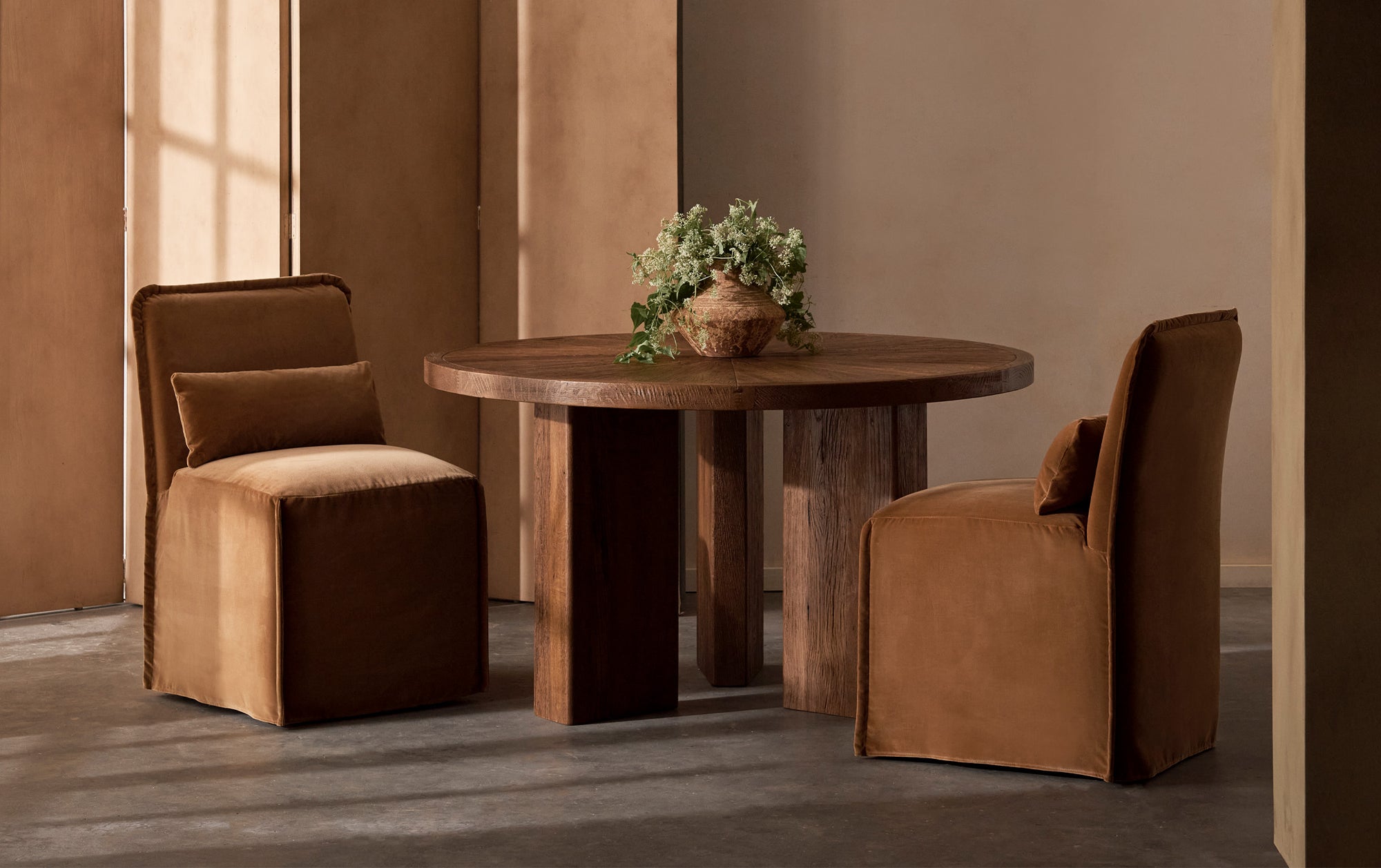 A set of Melo Armless Dining Chairs in Sugar Rush, a luscious caramel Washed Cotton Velvet, placed in a sunlit room at a Tango Round Dining Table in Heritage Oak, with a small vase of flowers as its centerpiece.