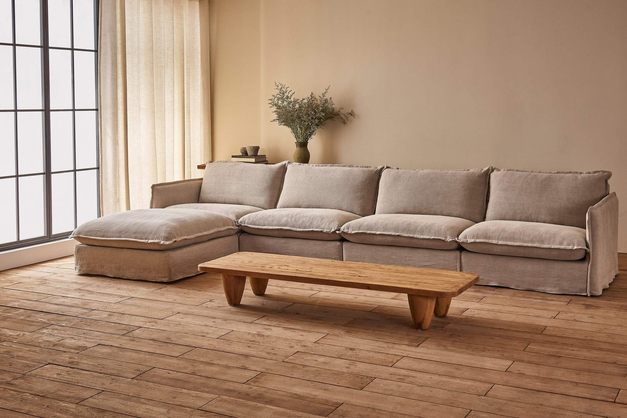 Neva 5-piece Chaise Sectional Sofa in Jasmine Rice, a light warm greige Medium Weight Linen, placed in a warmly lit room with a Theo Coffee Table, in front of a decorated console table