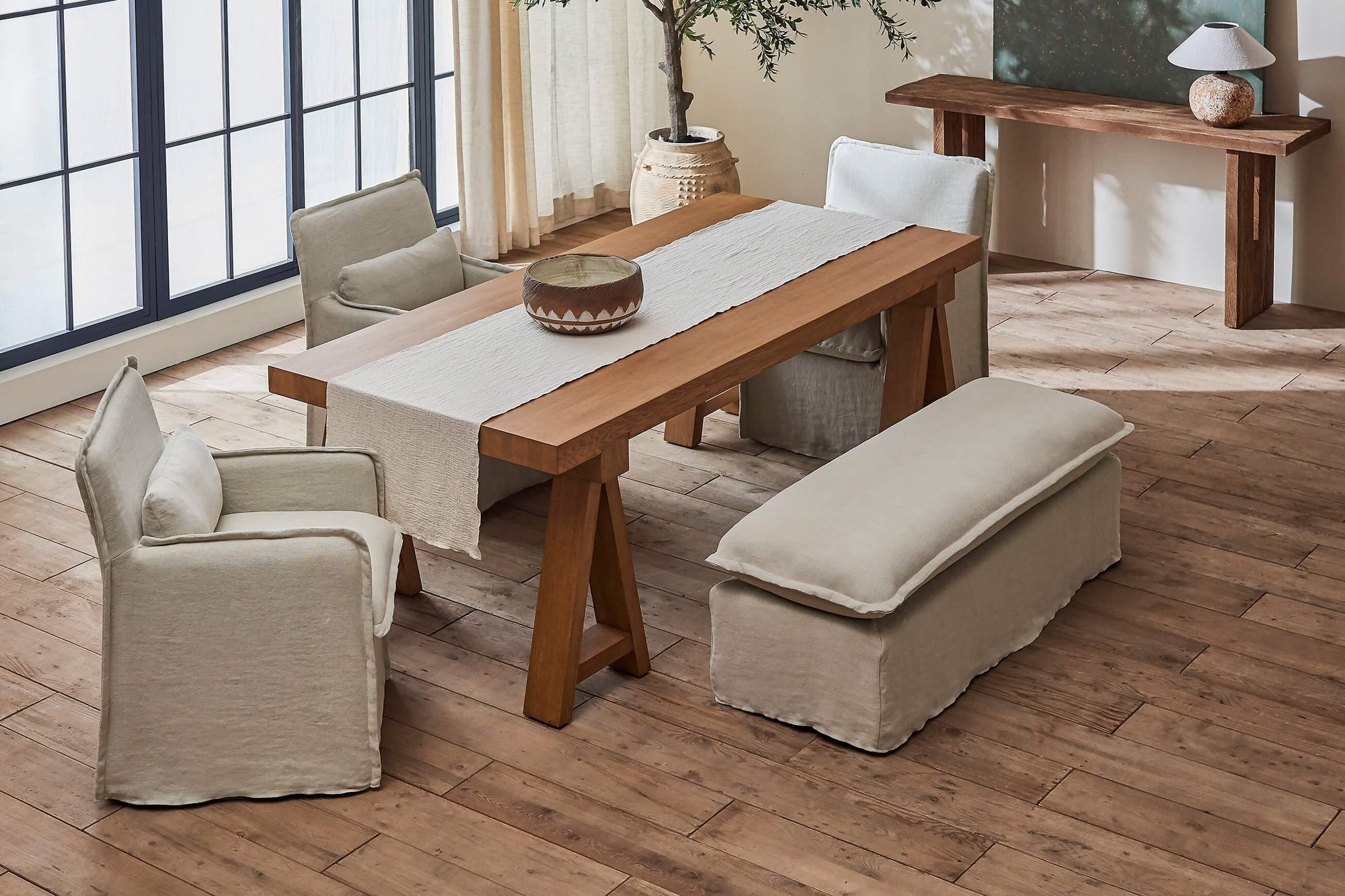 Neva Dining Bench in Warm Oatmeal, a light warm beige Medium Weight Linen, placed in a sunlit room surrounding the Rylance Dining Table and three Melo Dining Chairs