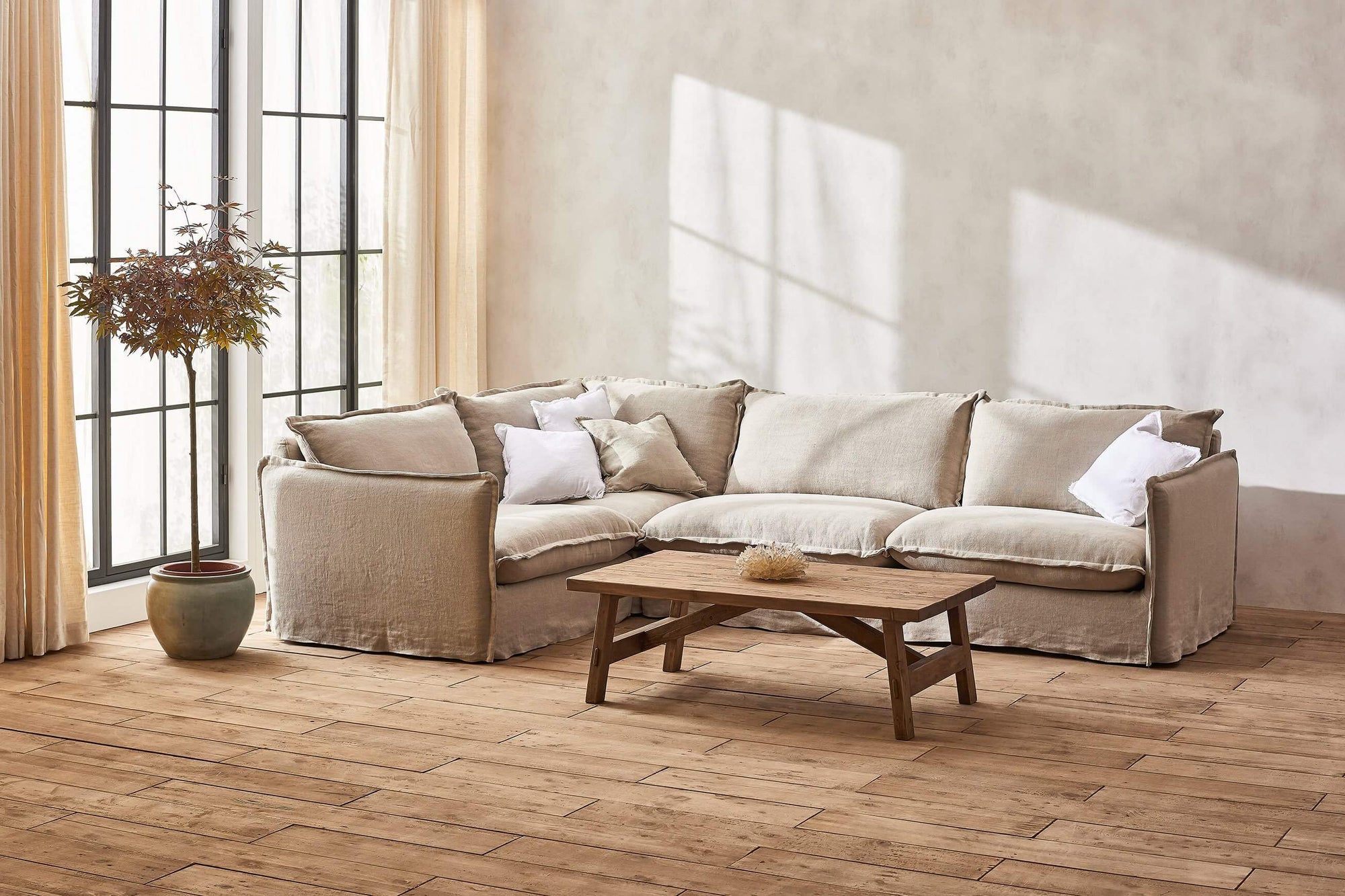 Neva L-Shape Sectional Sofa in Jasmine Rice, a light warm greige Medium Weight Linen, placed in a sunlit room with a coffee table and a small potted tree