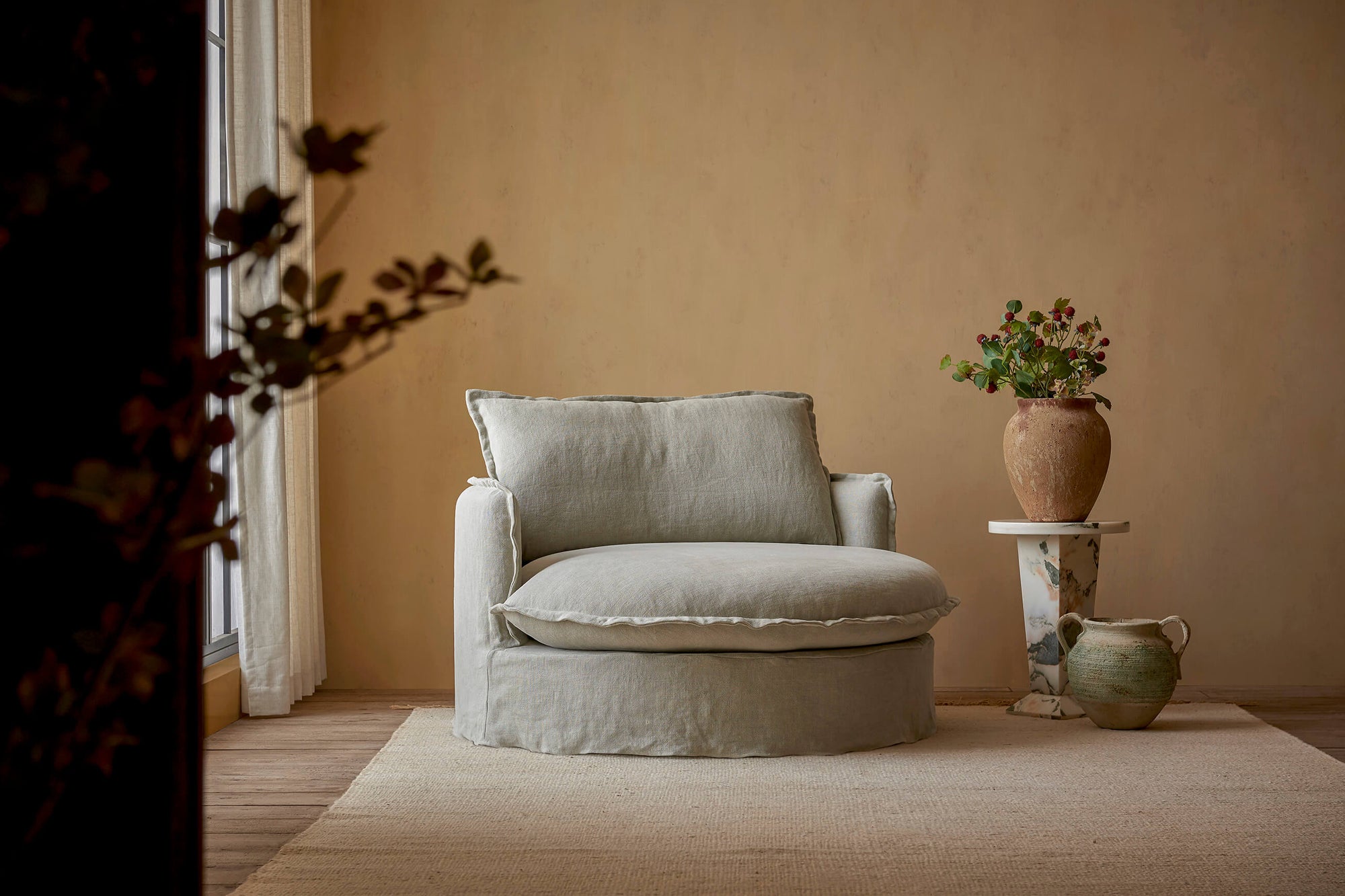 Neva Round Daybed in Jasmine Rice, a light warm greige Medium Weight Linen, placed in a sunlit room next to flowers in vases on a side table