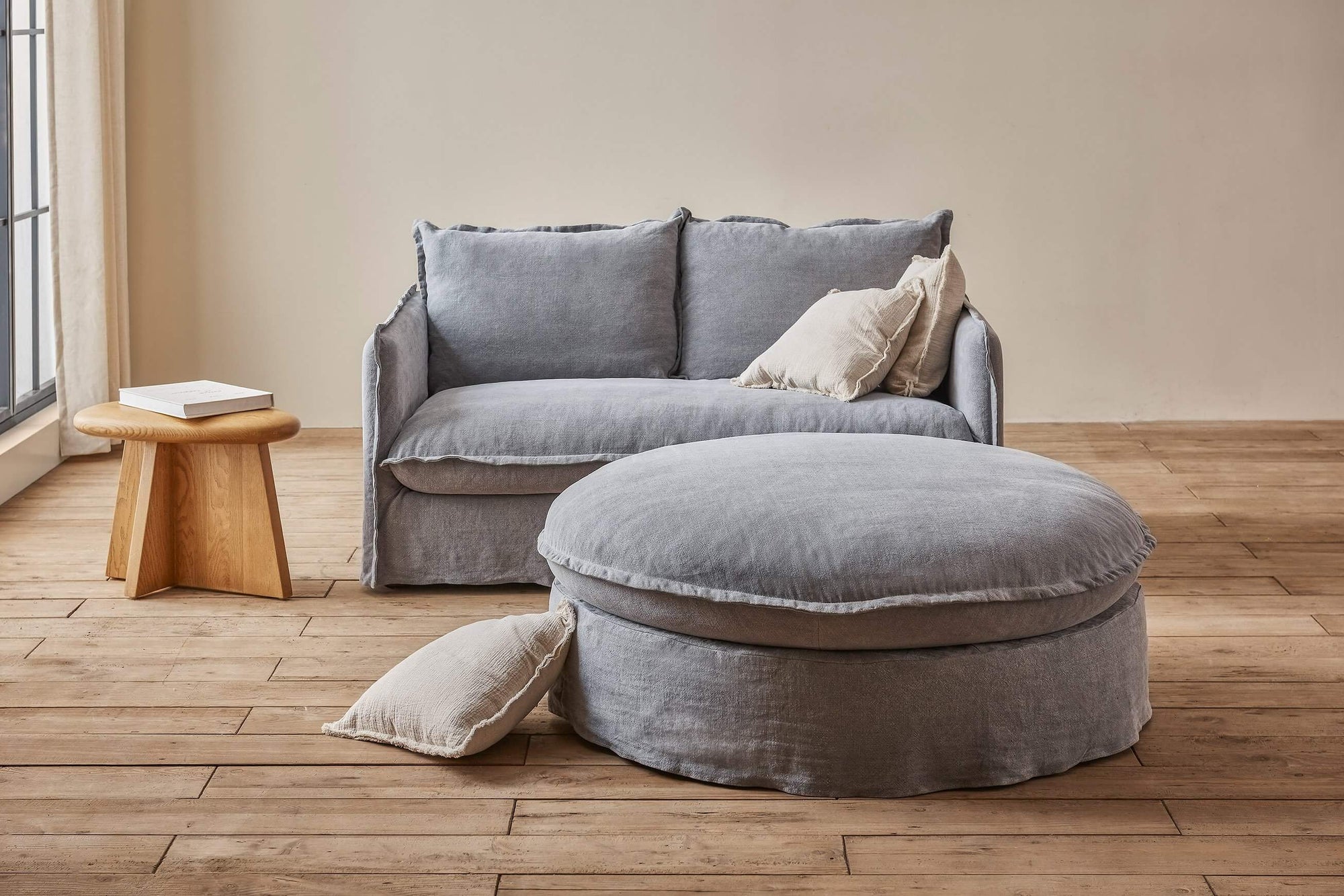 Neva Round Ottoman in Ink Cap, a medium cool grey Light Weight Linen, placed in fromt of a Neva Loveseat and an Amina side table.