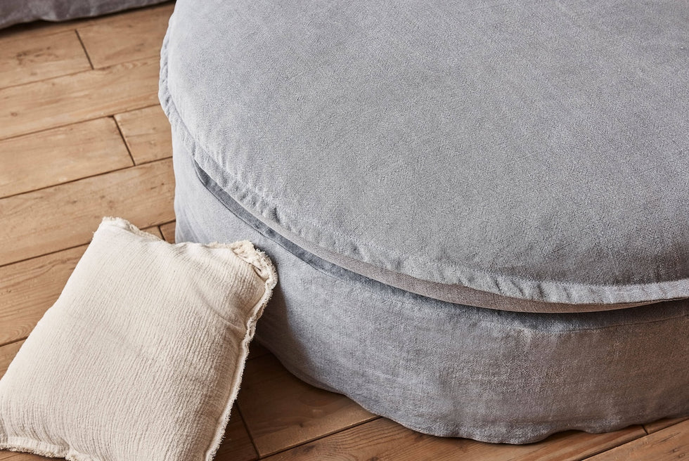 Close up of Neva Round Ottoman in Ink Cap, a medium cool grey Light Weight Linen, with a light beige throw pillow