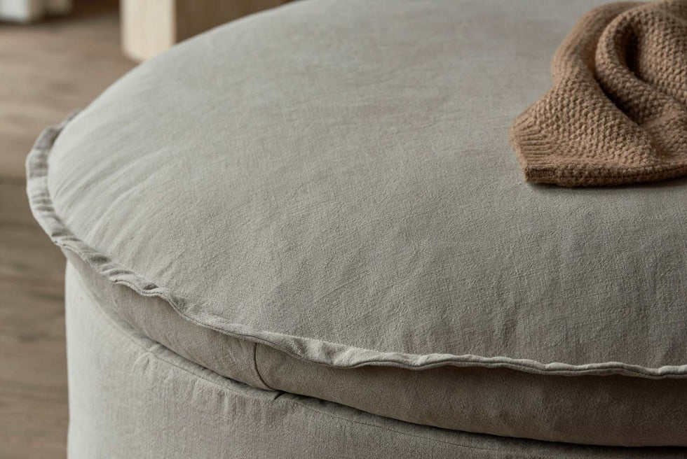 Close up of Neva Round Storage Ottoman in Oat Flour, a medium taupe Light Weight Linen, with a brown knit throw blanket