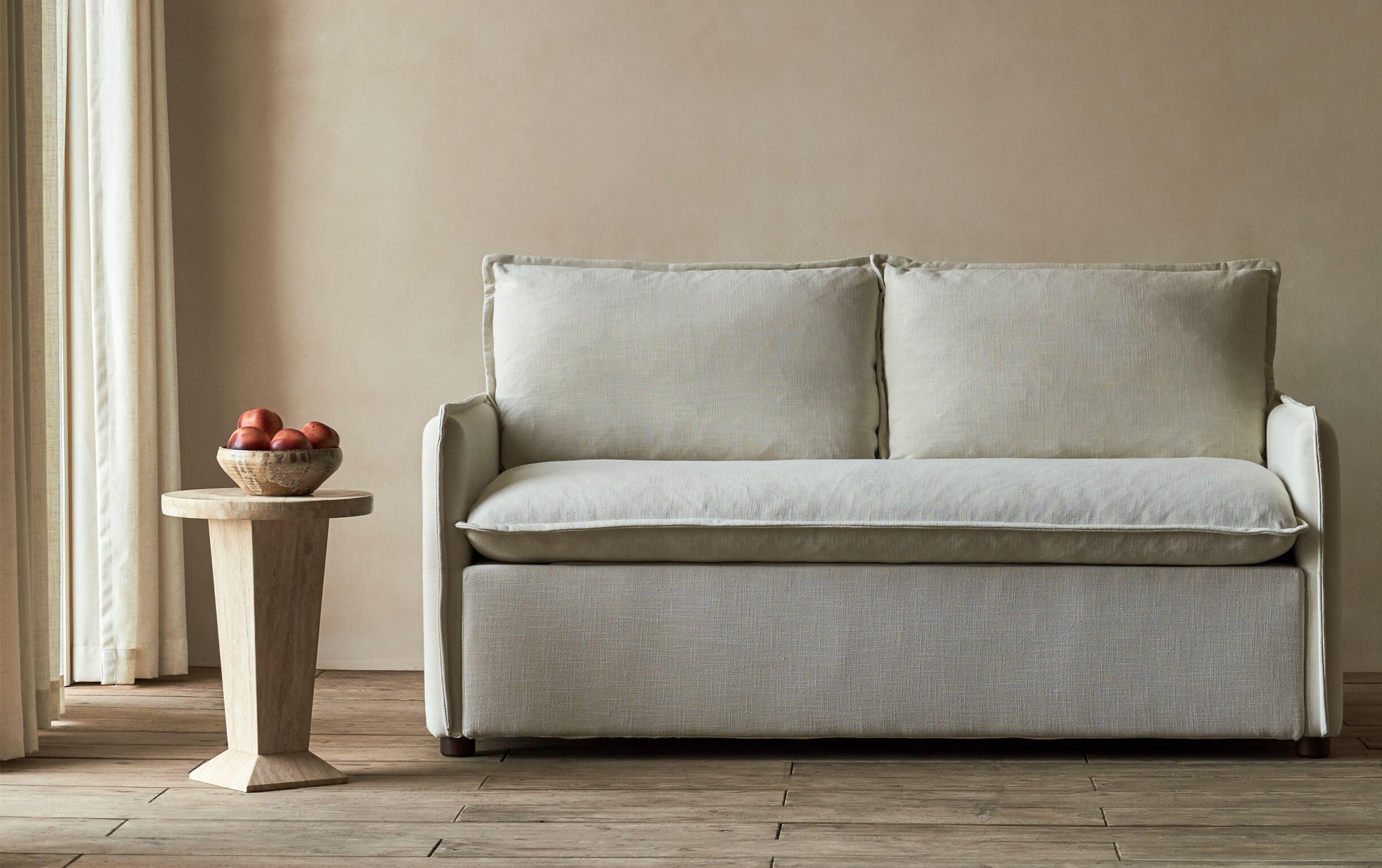 Neva Sleeper Sofa in Young Coconut, a powdery white Recycled Poly Linen, placed in a sunny room next to a stone side table holding a bowl of apples