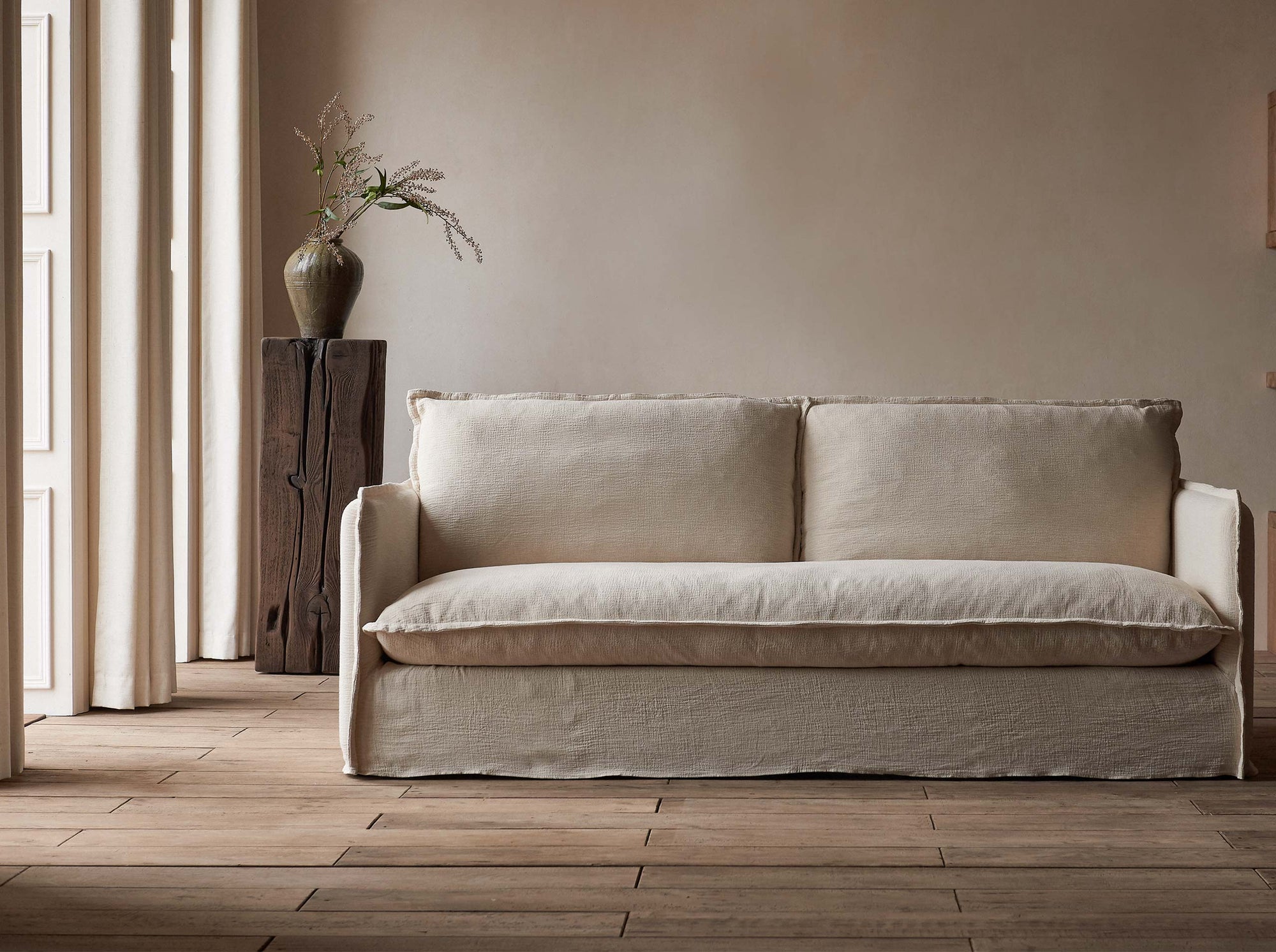 Neva 84" Sofa in Corn Silk, a light beige Washed Cotton Linen, placed in a softly lit room with a vase of flowers on a wooden pillar