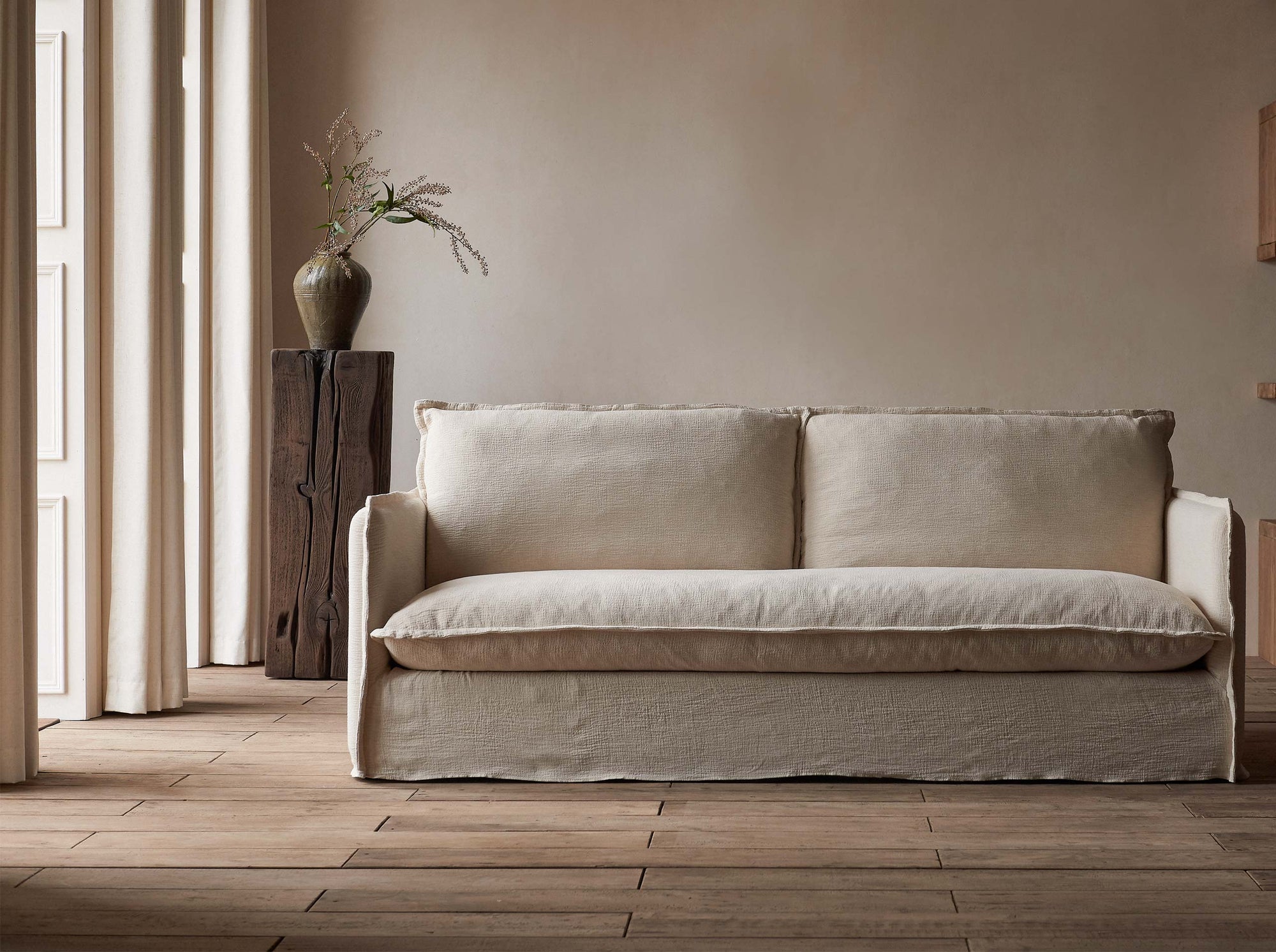 Neva 84" Sofa in Corn Silk, a light beige Washed Cotton Linen, in front of a wooden pedestal decorated with a vase and flowers