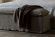 Slide #7 Close up of Neva Storage Bench in Jasmine Rice, a light warm greige Medium Weight Linen, with a brown throw blanket, placed at the end of a Neva King Bed