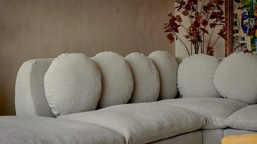 Close up of Olea L-Shape Sectional Sofa in Jasmine Rice, a light warm greige Medium Weight Linen, placed in a room decorated with art and a plant