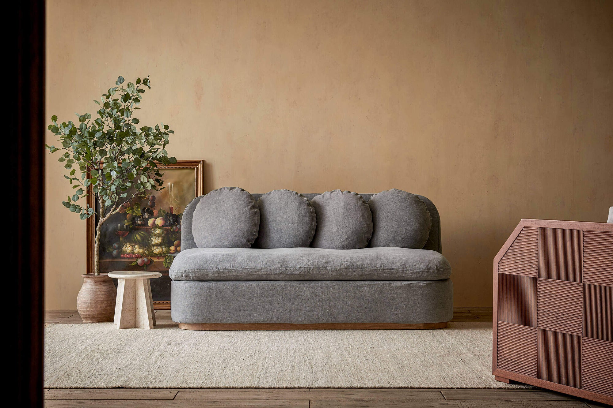 Olea 72" Sofa in Ink Cap, a medium cool grey Light Weight Linen, next to a side table, a framed painting, and a large potted plant