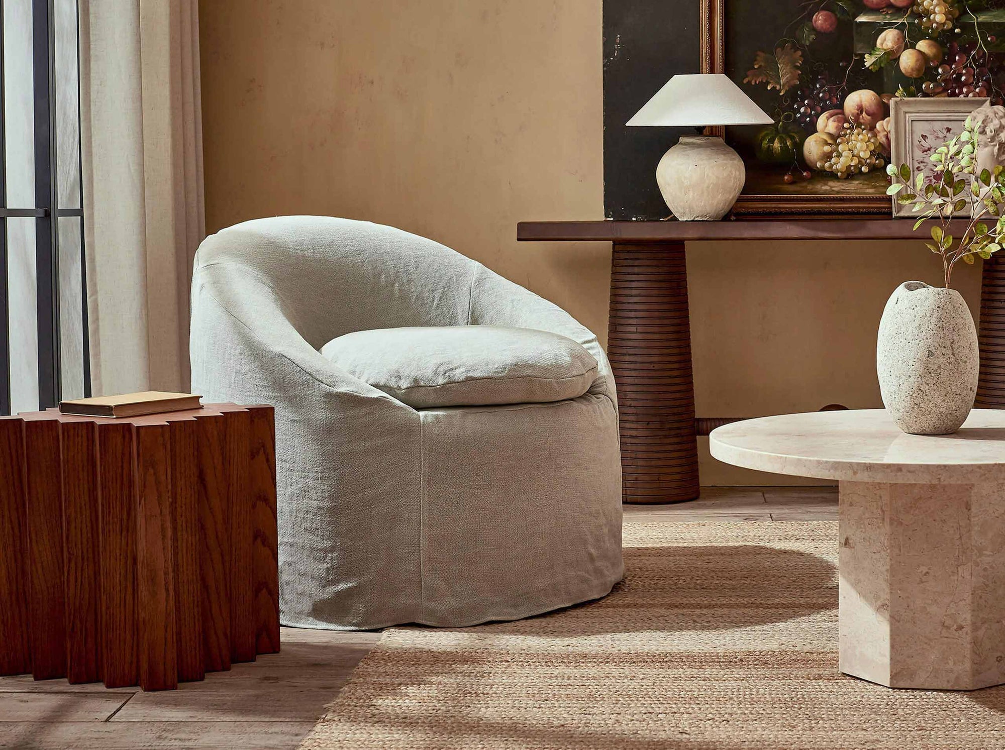 Ozzi Chair in Jasmine Rice, a light warm greige Medium Weight Linen, placed in a decorated room with a wooden side table and round stone coffee table