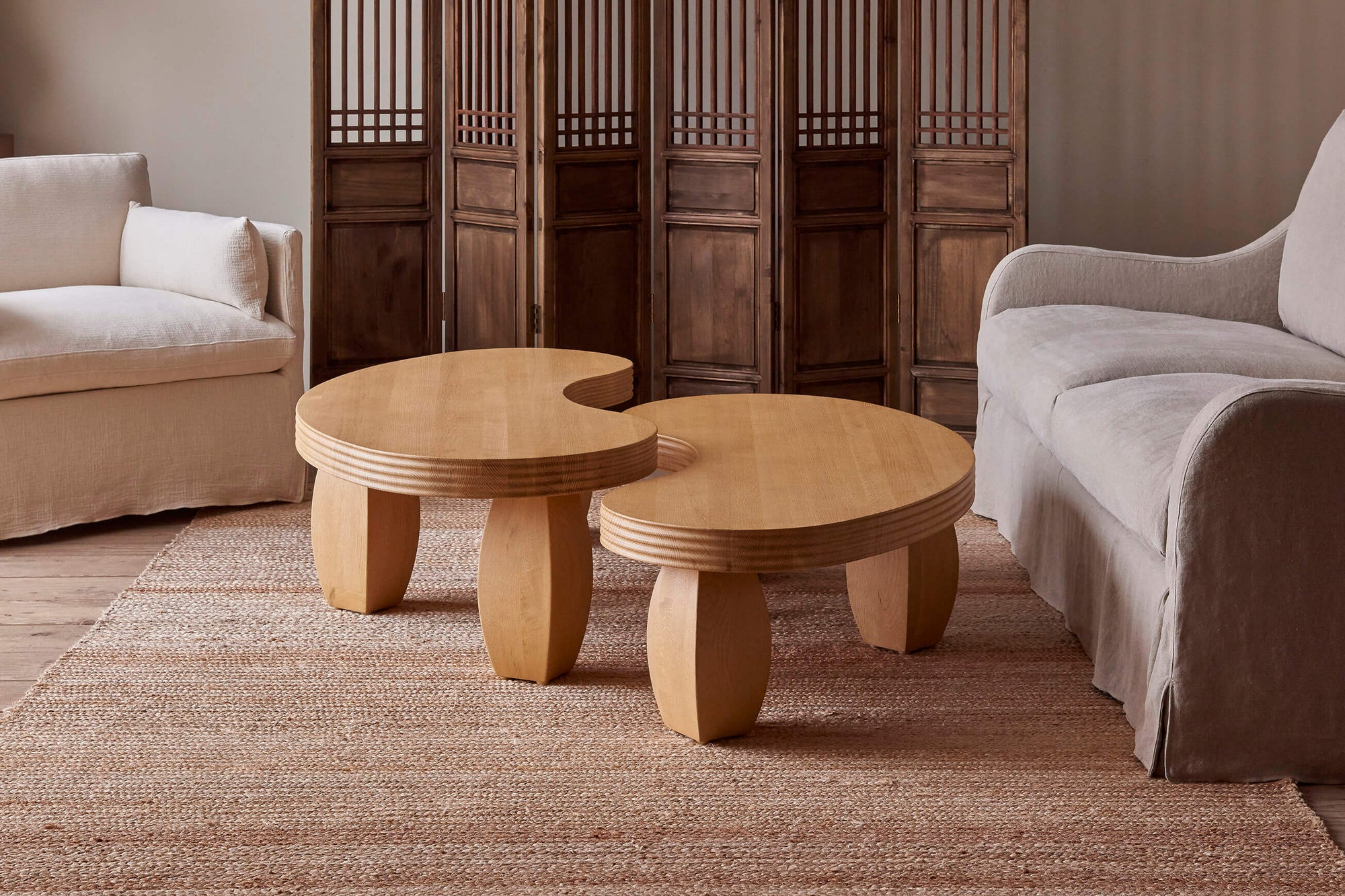 A Short and Tall Pisces Coffee Table in Pale Oak placed next to each other in front of a wooden folding screen, between the Gabriel Chair and Esme Sofa