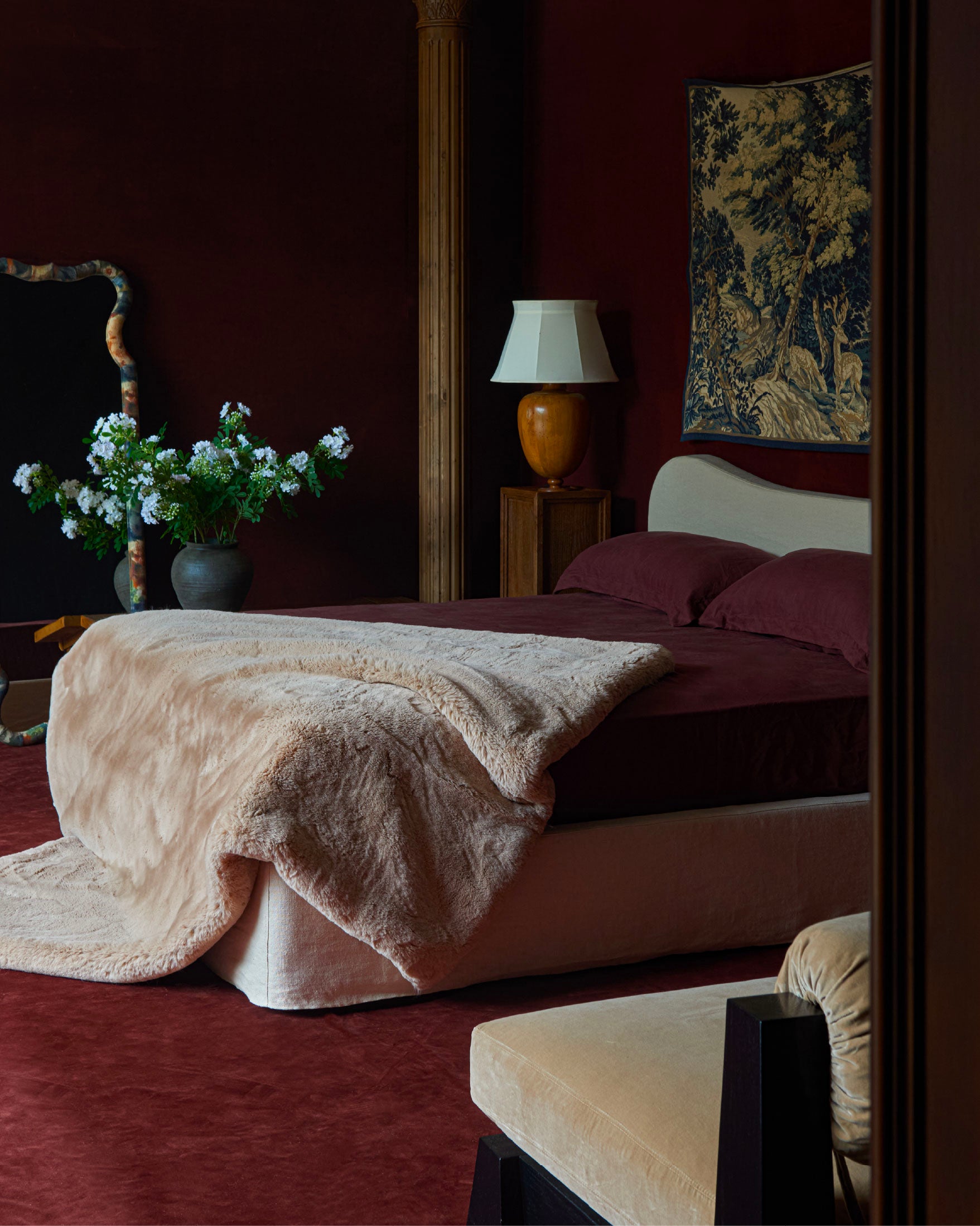 A luxurious bedroom scene featuring a sixpenny recycled faux fur pampas flow blanket draped over the foot. The room has a matching deep burgundy carpet and wall, decorated with a vintage-style tapestry, a wooden table lamp, and a vase of white flowers.