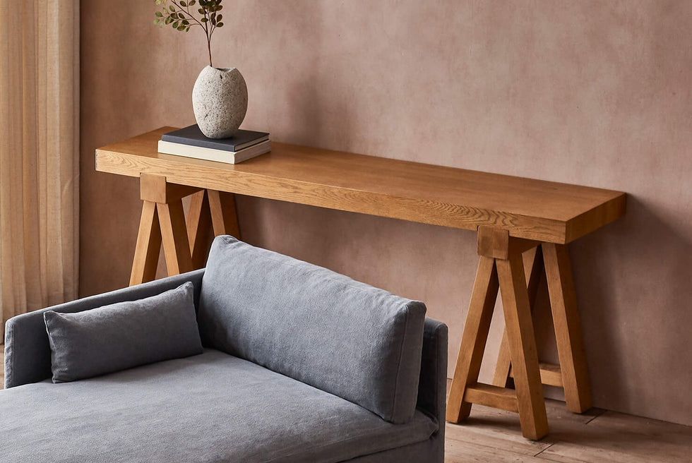 Rylance Console Table in Tinted Oak, decorated with books and a stone vase, placed against a wall behind a Gabriel Daybed in Ink Cap.