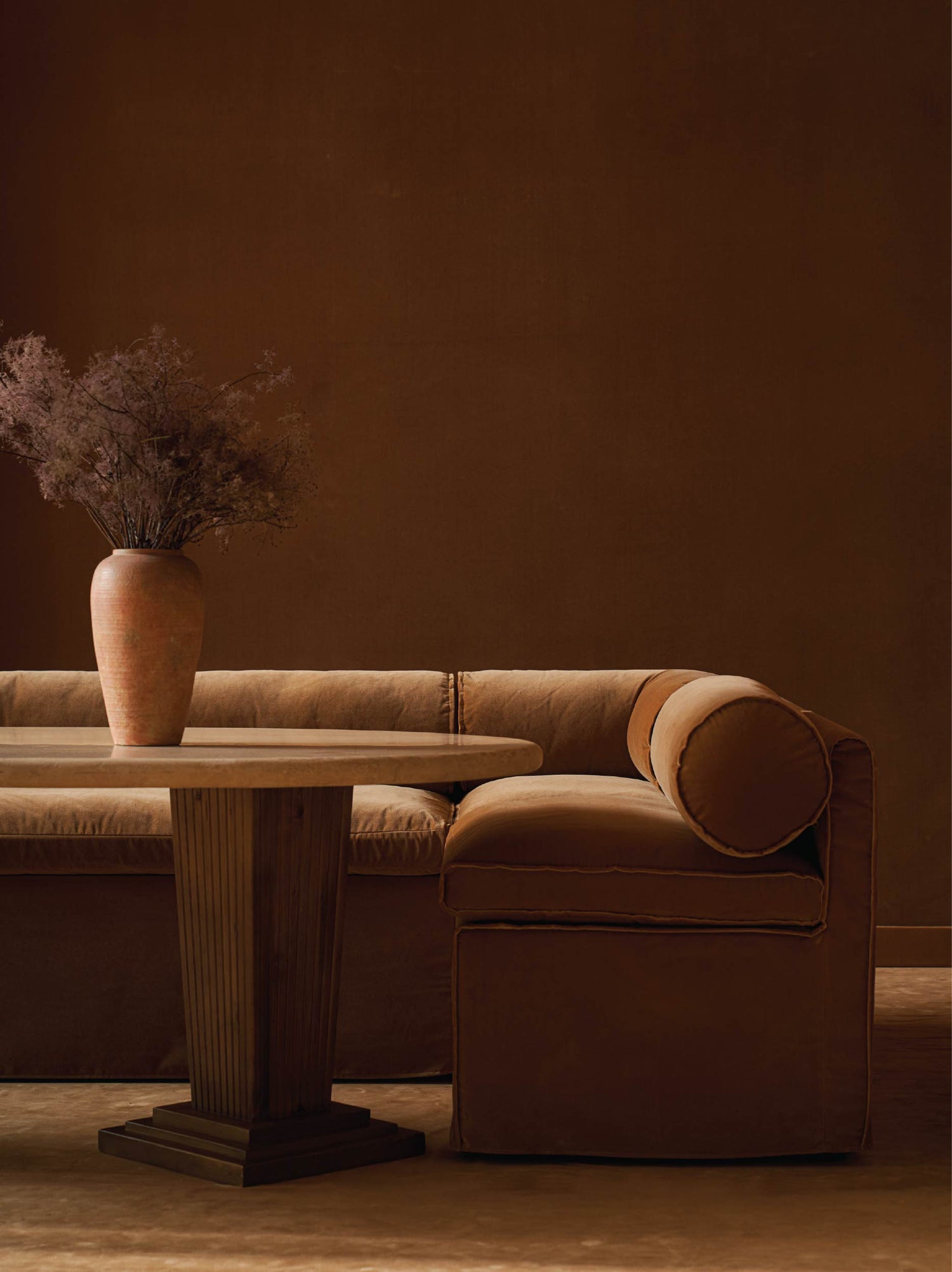 sixpenny camino banquette sofa and Pisces Coffee Table with a vase of dried flowers against a brown wall.