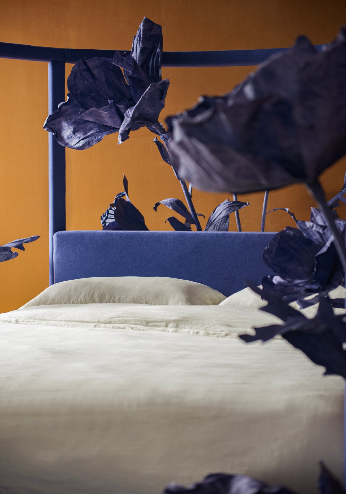 A close-up of a sixpenny florette round bed in fountainhead with cream-colored bedding and a blue upholstered headboard, set against a warm orange wall with dark blue metal bed frames and large dark purple decorative leaves in the foreground.