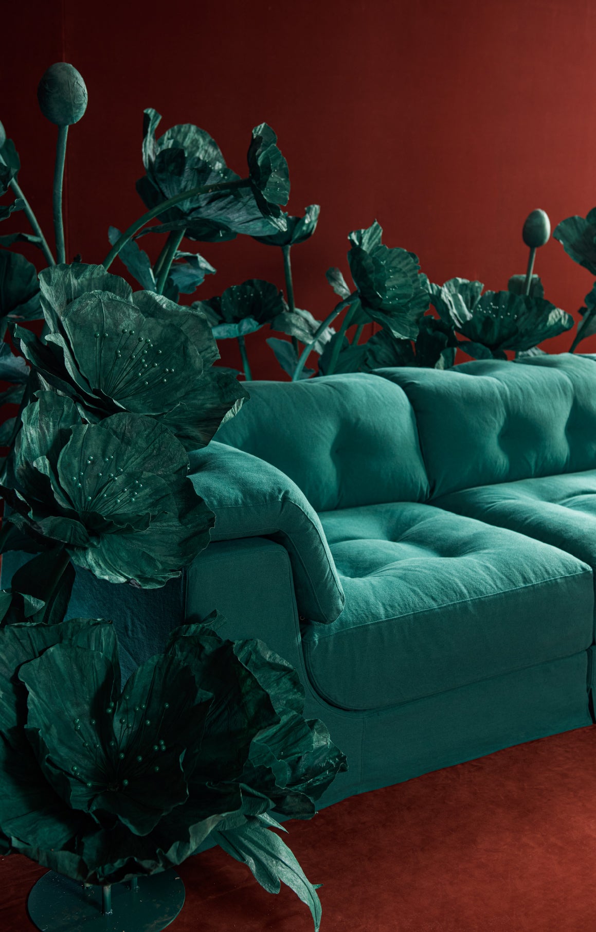 A sixpenny Mo sectional sofa in Ivy League, styled in a room with a matching dark red wall and floor, surrounded by oversized dark green decorative leaf-like plants.