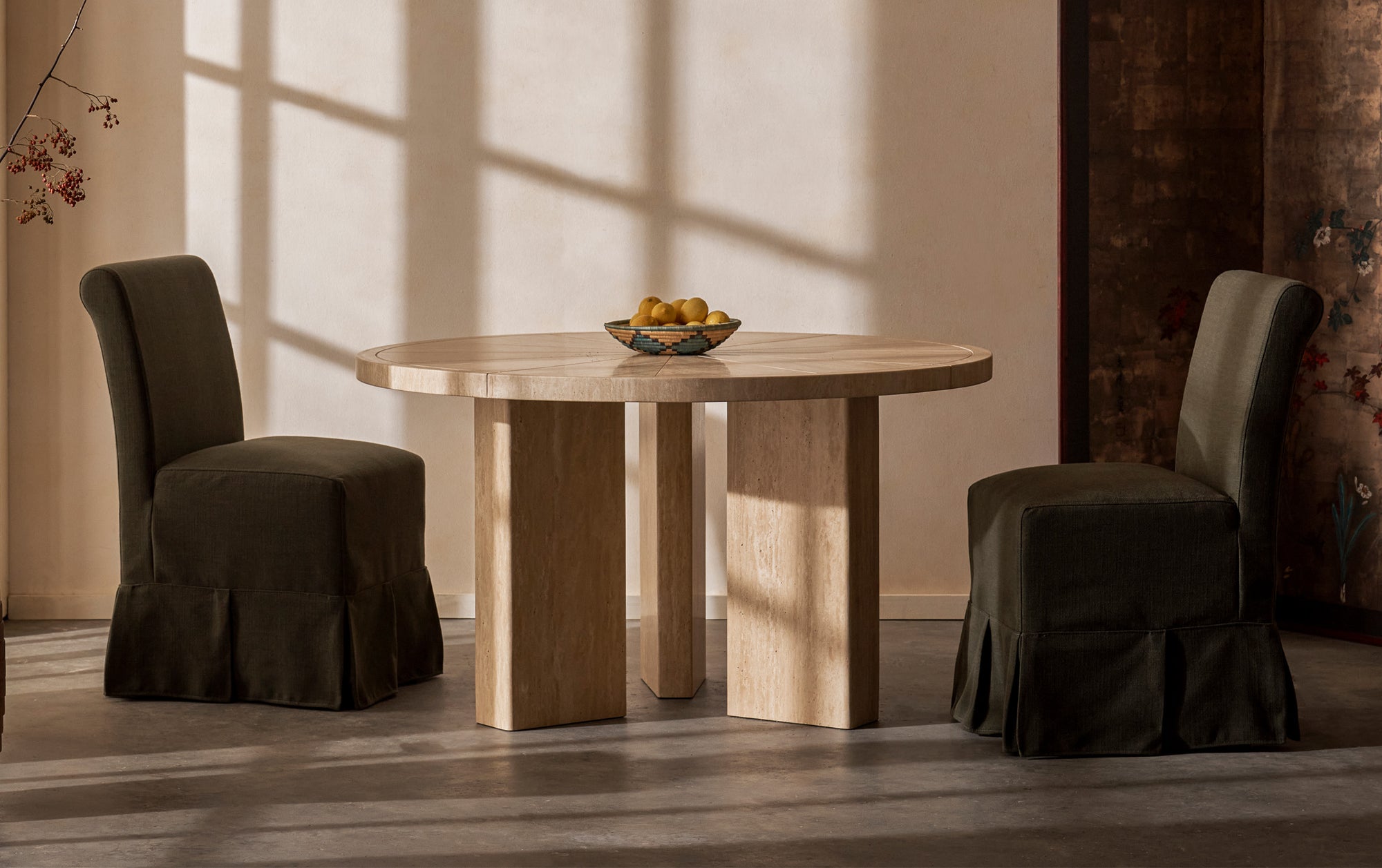 A set of Dalia Armless Dining Chairs in New Leaf at a Tango Round Dining Table in Turkish Galata Travertine in a warm beige with gold and pink undertones-- various veining, color variations, and pitting throughout-- placed in a warmly lit room and decorated with a woven bowl of lemons.