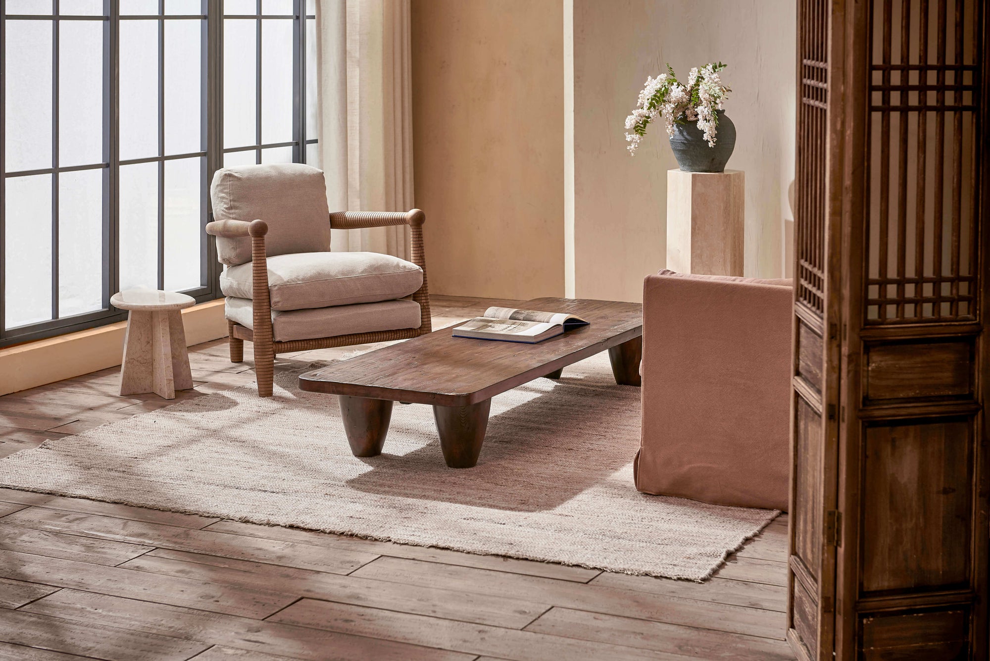 Theo Coffee Table in Dusky Pine, a deep, rich, molasses colored 100% Chinese pine, with an open book on the tabletop, placed in front of a large window with a Gio Chair