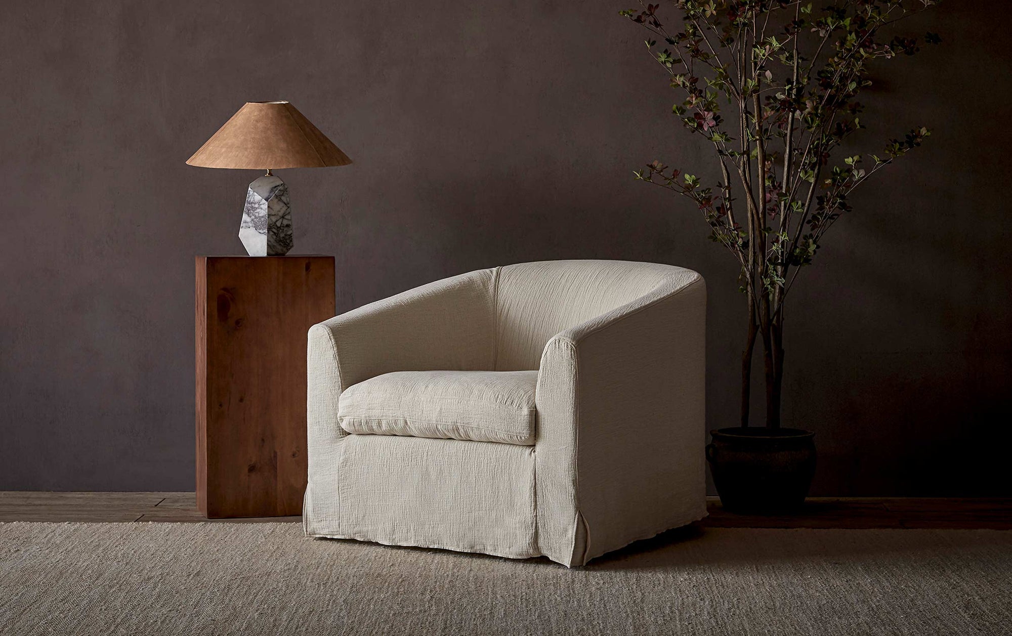 Ziki Chair in Corn Silk, a light beige Washed Cotton Linen, placed in a room between a potted tree and a table lamp on a side table