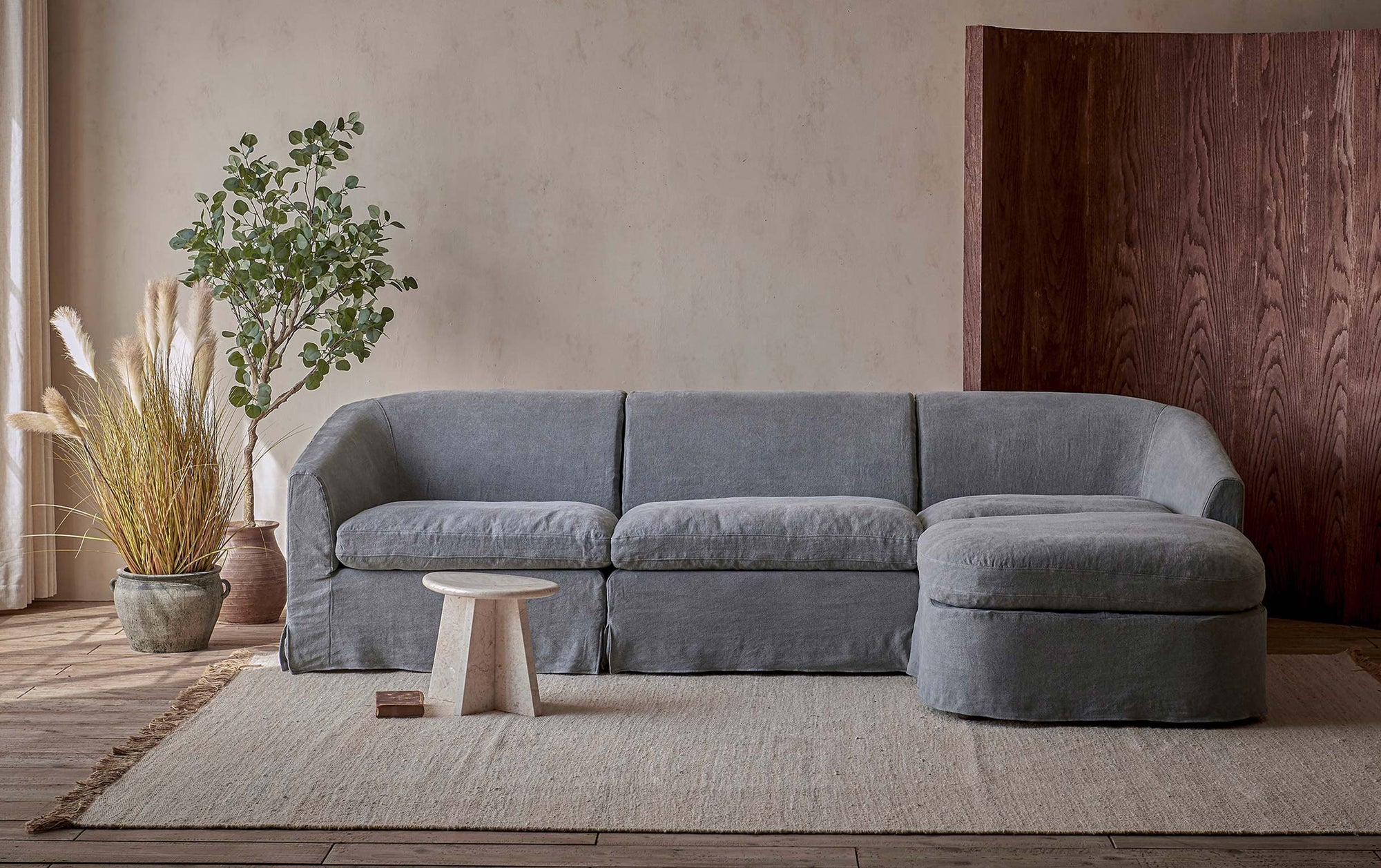 Ziki 4-piece Chaise Sectional Sofa in Ink Cap, a medium cool grey Light Weight Linen, placed in a room with plants and a stool