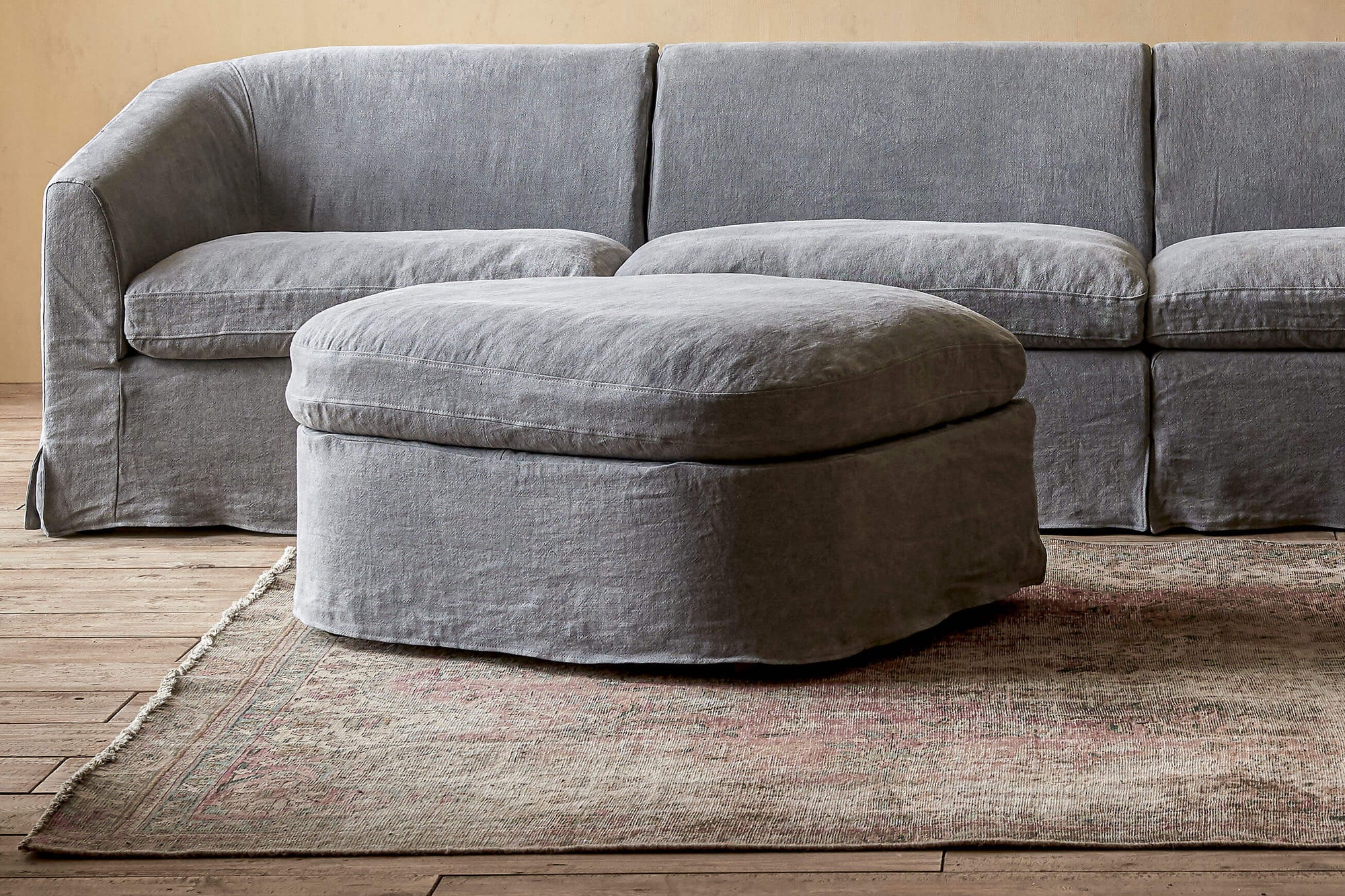 Ziki Sectional Ottoman in Ink Cap, a medium cool grey Light Weight Linen, placed on a rug in front of the Ziki Sectional Sofa