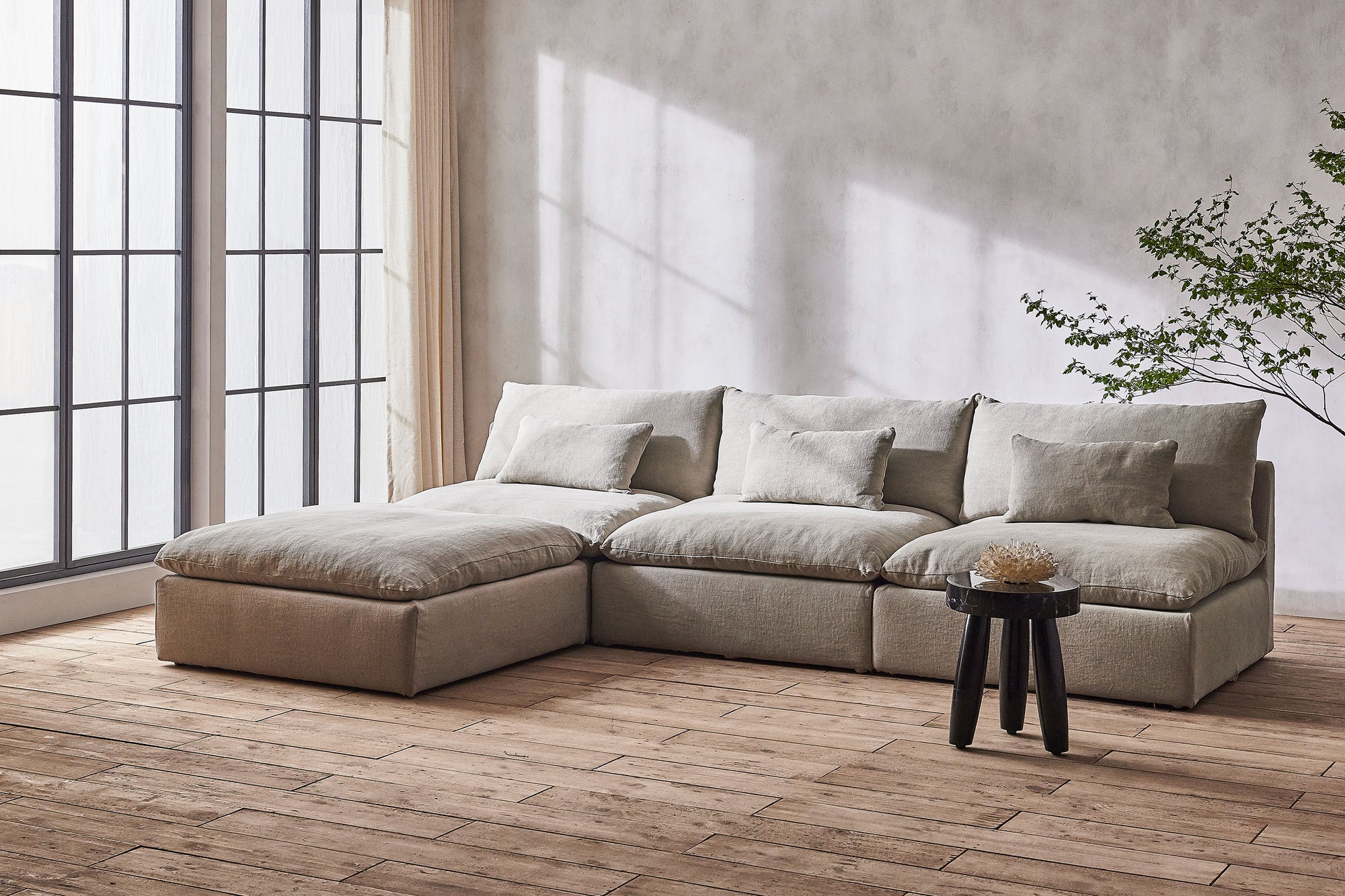 Aria 4-piece Chaise Sectional Sofa in Jasmine Rice, a light warm greige Medium Weight Linen, placed in a sunlit room next to a stool