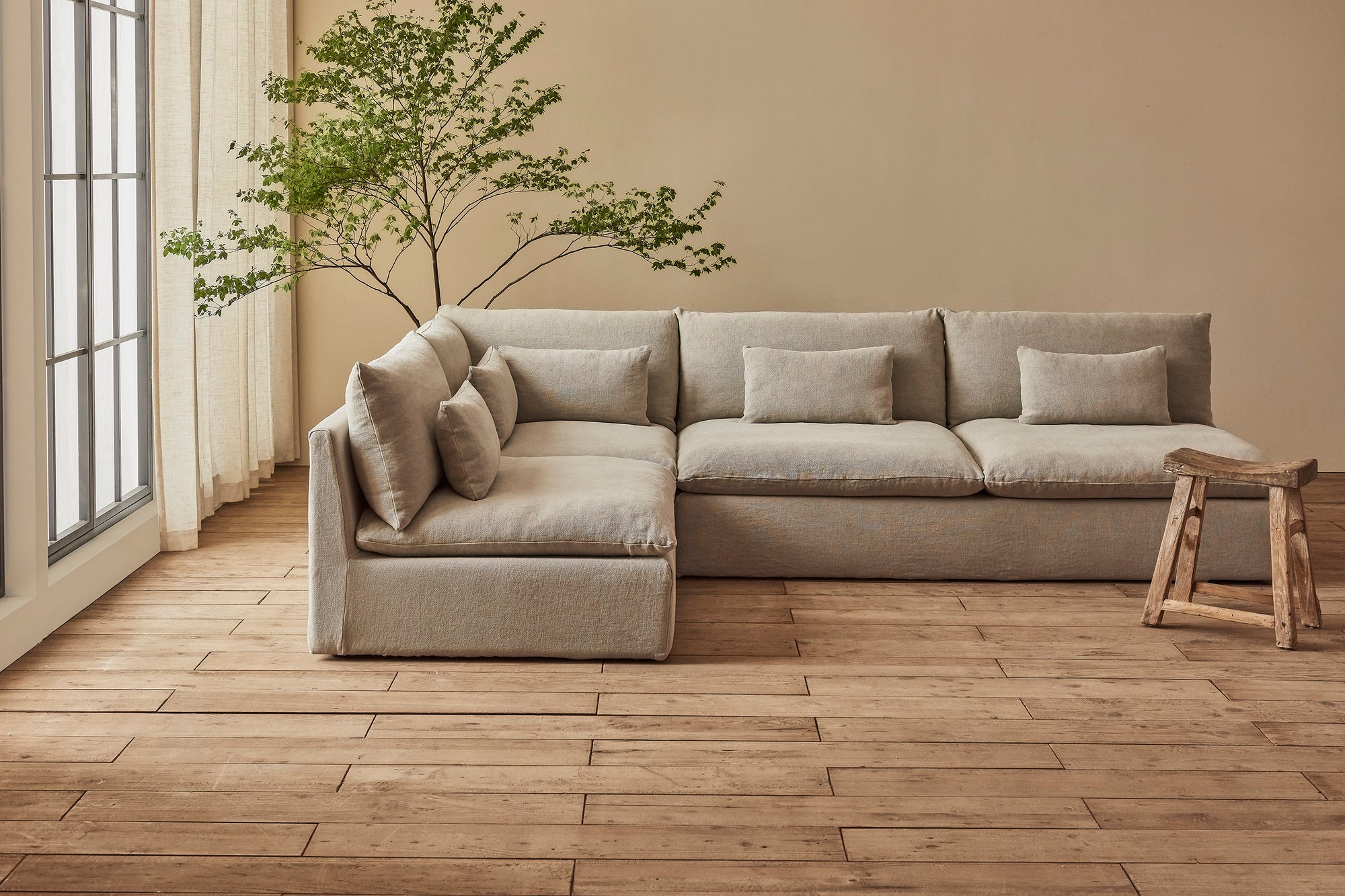 Aria Long L-Shape Sectional Sofa in Jasmine Rice, a light warm greige Medium Weight Linen, placed in a sunlit room between a potted tree and a stool