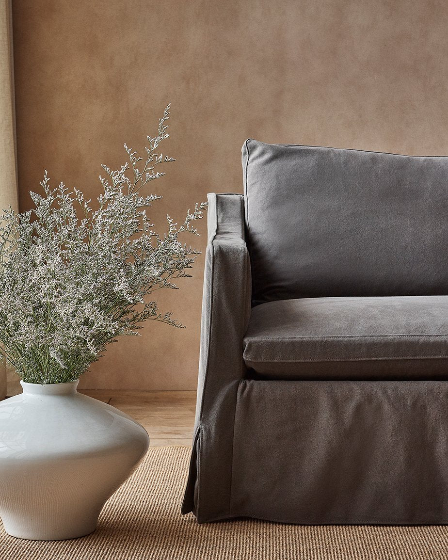 Amelia Sofa in Moon Dust, a dark charcoal grey Cotton Canvas, placed in a room next to a flowering plant in a vase on the floor