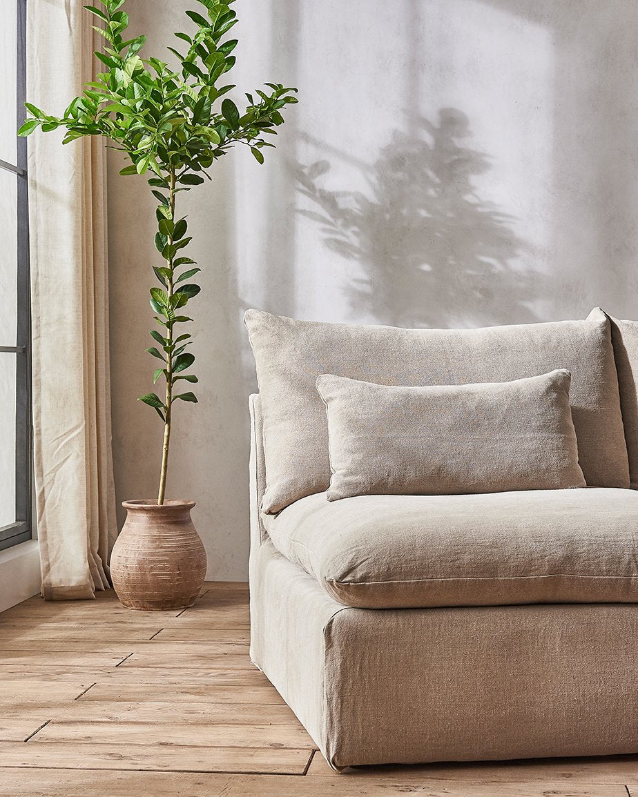 Aria Sofa in Oat Flour, a medium taupe Light Weight Linen, placed in a sunlit room in front of a potted tree