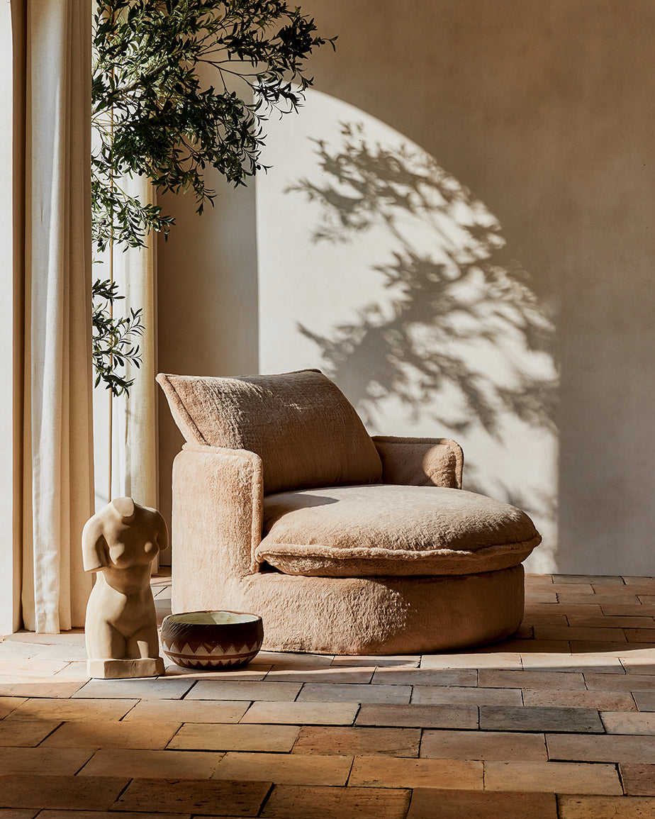 Neva Round Daybed in Pampas Flow, a light tan brown Recycled Faux Fur, placed in a sunlit room next to a statue and vase on the floor