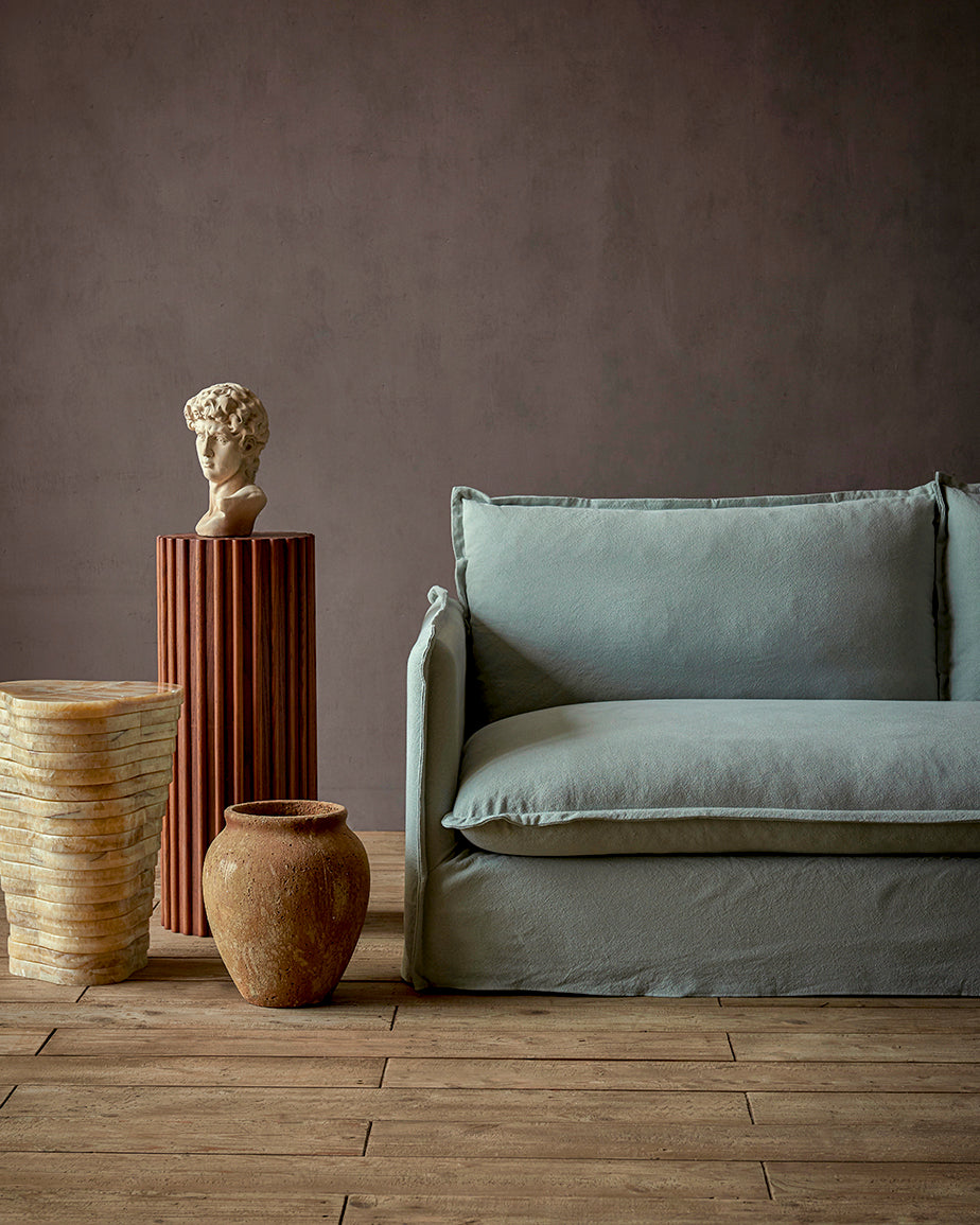 Neva Sofa in Hello Aloe, a pale green Thread-Dyed Cotton Linen, placed in a room next to a decorative pillar and vases