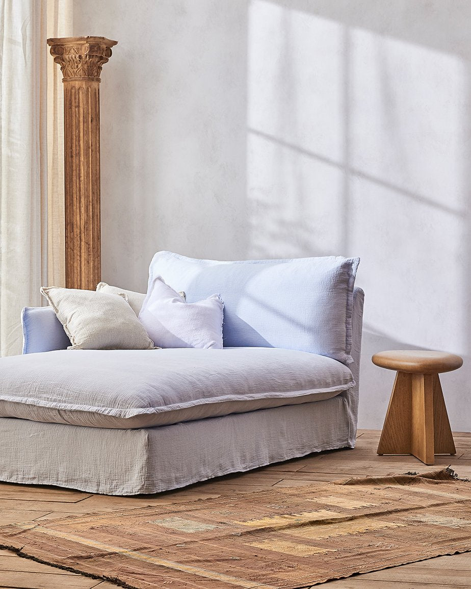 Neva Daybed in Morning Glory, a pale blue Washed Cotton Linen, placed in a sunlit room between a pillar and a stool 